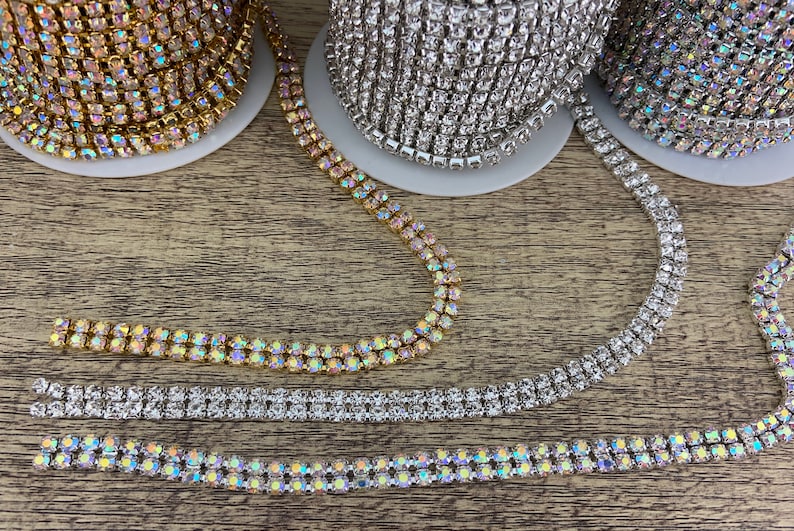 K&ouml;nnte beinhalten: Drei Spulen mit Strassbesatz, eine gold, eine silber und eine regenbogenfarben. Der Besatz ist zweireihig und liegt auf einer Holzoberfl&auml;che.