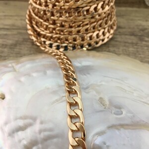 May include: A gold curb chain necklace with a thick, flat link design. The chain is lying on a white, textured surface.