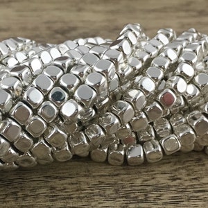 May include: Close-up of a collection of shiny, silver cube beads. The beads are strung together, forming a flexible strand. The beads have rounded edges and reflect light, creating a bright, metallic appearance. Ideal for jewelry making.