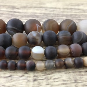 May include: A strand of brown agate beads with a variety of patterns and shades. The beads are round and smooth, and they are arranged in a row on a wooden surface.