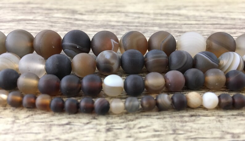 May include: A strand of brown agate beads with a variety of patterns and shades. The beads are round and smooth, and they are arranged in a row on a wooden surface.