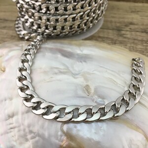 May include: A silver curb chain necklace with a thick, flat link design. The chain is lying on a white, pearlescent surface.