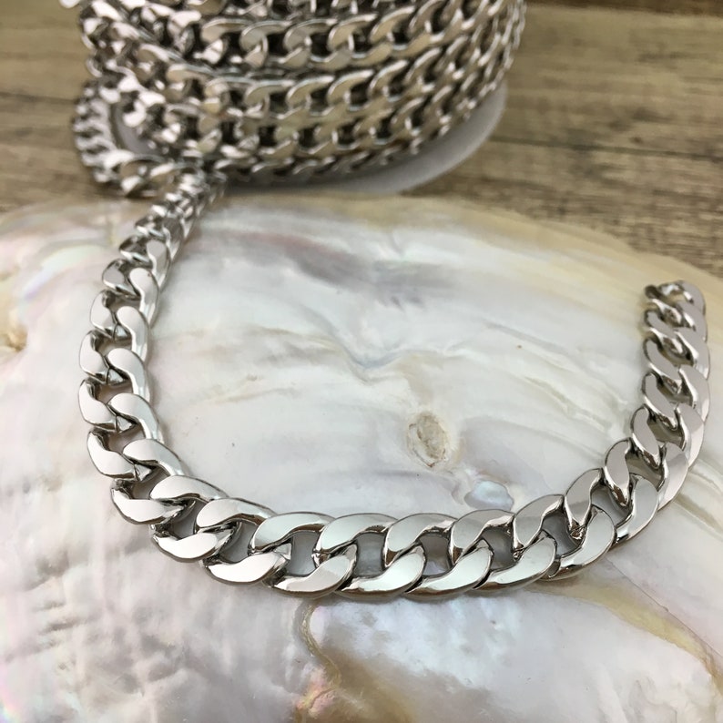 May include: A silver curb chain necklace with a thick, flat link design. The chain is lying on a white, pearlescent surface.
