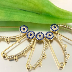 May include: Five gold safety pins with blue evil eye charms and clear rhinestones. The pins are arranged in a fan shape.