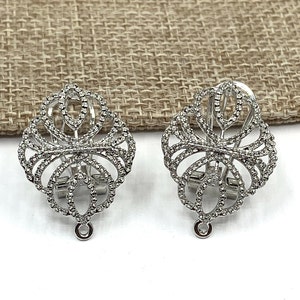 May include: A pair of silver tone clip-on earrings with a delicate, intricate design. The earrings are made of metal and have a small loop at the bottom.