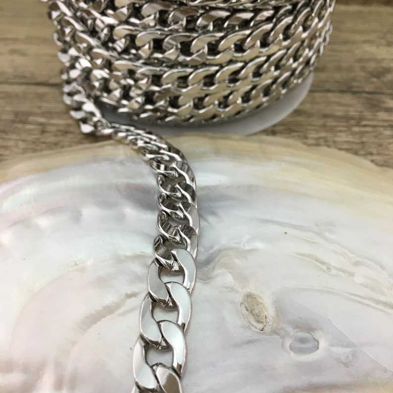 May include: Silver curb chain trim, a close-up of the chain links. The chain is lying on a white, iridescent surface.