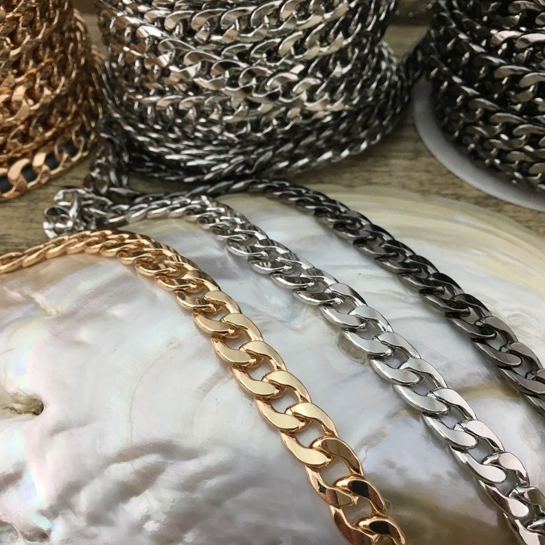May include: Three curb chains in gold, silver, and black metal. The chains are laid out on a white, iridescent surface.