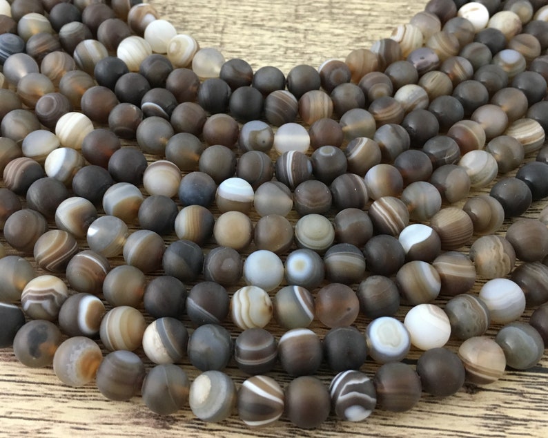 May include: A strand of brown agate beads with white and gray banding. The beads are round and smooth, and they are arranged in a loose pile.