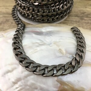 May include: A close-up of a black metal chain with a curb link design. The chain is lying on a white surface.