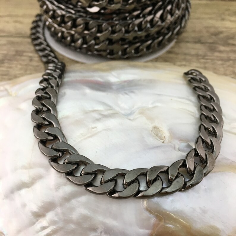 May include: A close-up of a black metal chain with a curb link design. The chain is lying on a white surface.