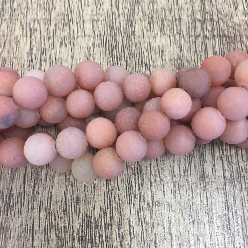 May include: A strand of smooth, round, light pink and peach colored agate beads. The beads are matte and have a slightly textured surface.