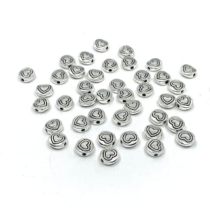May include: A scattering of silver-toned metal beads with a heart design. The beads have a black outline and a small hole for stringing.