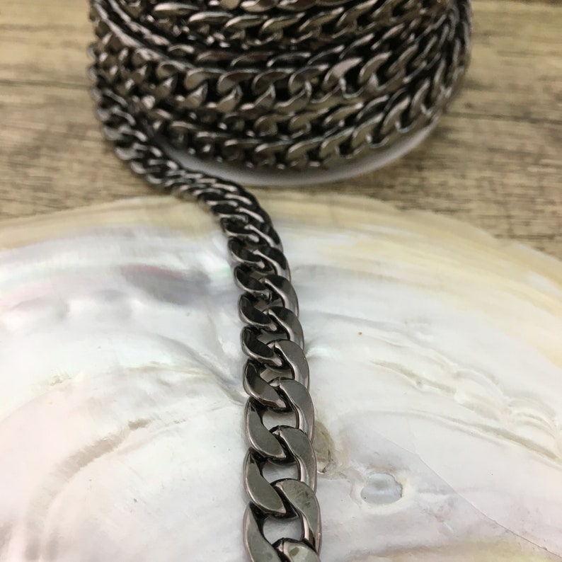 May include: A close-up of a black metal chain with a curb link design. The chain is lying on a white surface.