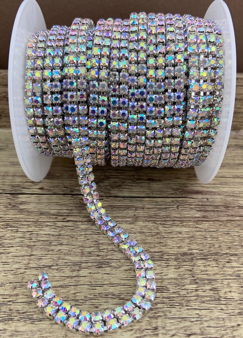 K&ouml;nnte beinhalten: Eine Spule mit silberner Strass-Kettenborte. Die Kette besteht aus kleinen, quadratischen Strasssteinen, die in einer silbernen Metallfassung gefasst sind. Die Kette ist schillernd und hat einen Regenbogeneffekt.