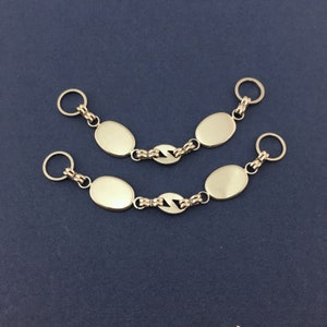 Può includere: Due accessori a catena color argento, ciascuno con tre maglie ovali e un portachiavi a ciascuna estremità. Le maglie sono collegate da una serie di maglie stilizzate a centro aperto. Lo sfondo è blu scuro.