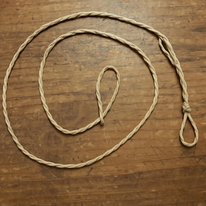 Può includere: Una corda beige intrecciata con un anello e un nodo all'estremità, appoggiata su una superficie di legno. La corda è avvolta in un motivo circolare, con un anello più piccolo al centro. La trama della corda è visibile.