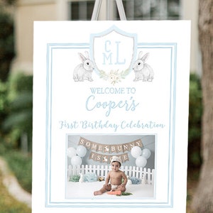 May include: A blue and white welcome sign for a first birthday party. The sign says "Welcome to Cooper's First Birthday Celebration". There are two bunnies on the sign and a photo of a baby boy in a white shirt and a blue hat. The photo shows the baby sitting in front of a banner that says "Some Bunny Is One".