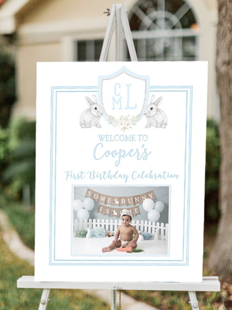 May include: A blue and white welcome sign for a first birthday party. The sign says "Welcome to Cooper's First Birthday Celebration". There are two bunnies on the sign and a photo of a baby boy in a white shirt and a blue hat. The photo shows the baby sitting in front of a banner that says "Some Bunny Is One".