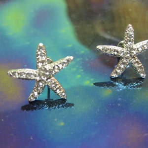 May include: A pair of silver starfish earrings with clear rhinestones. The earrings are on a blue and green iridescent background.