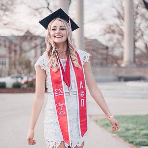 May include: A woman wearing a white lace dress and a red and white graduation sash with the text "Class of 2018" and Greek letters. She is wearing a black graduation cap and smiling.