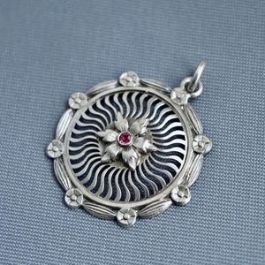 Könnte beinhalten: Silberanhänger mit floralem Design. Der Anhänger hat eine zentrale Blume mit einem roten Edelstein und ist von einem kreisförmigen Muster aus wellenförmigen Linien umgeben. Kleine Blumenakzente umranden den äußeren Rand.