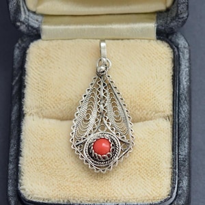 May include: A silver filigree pendant with a teardrop shape, featuring a small, round, red coral stone at its center. The pendant is displayed inside a vintage jewelry box with a cream-colored interior.