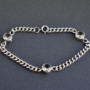 Puede incluir: Una pulsera de plata con un diseño de cadena de eslabones. La pulsera presenta tres piedras preciosas redondas y transparentes espaciadas uniformemente a lo largo de la cadena. El cierre es de diseño simple y circular.