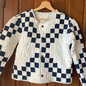 Könnte beinhalten: Gesteppte Jacke mit weißem Hintergrund und marineblauem Schachbrettmuster. Die Jacke hat lange Ärmel und eine Knopfleiste vorne. Die Jacke hängt an einem Holzbügel.