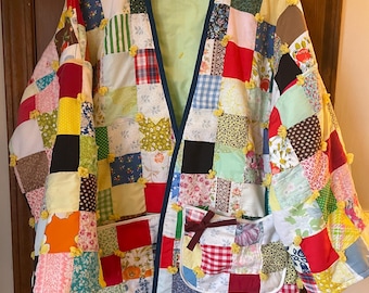 Puede incluir: Una chaqueta de patchwork con una variedad de cuadrados de tela coloridos. La chaqueta tiene un forro verde claro, ribete azul oscuro y dos bolsillos. Pompones amarillos están cosidos en las costuras. La chaqueta está colgada en una percha de madera.