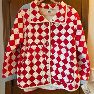 May include: A quilted jacket featuring a red and white diamond checkerboard pattern with small floral accents. The jacket has a wide collar, long sleeves with light blue cuffs, two large front patch pockets, and two blue buttons down the center front.