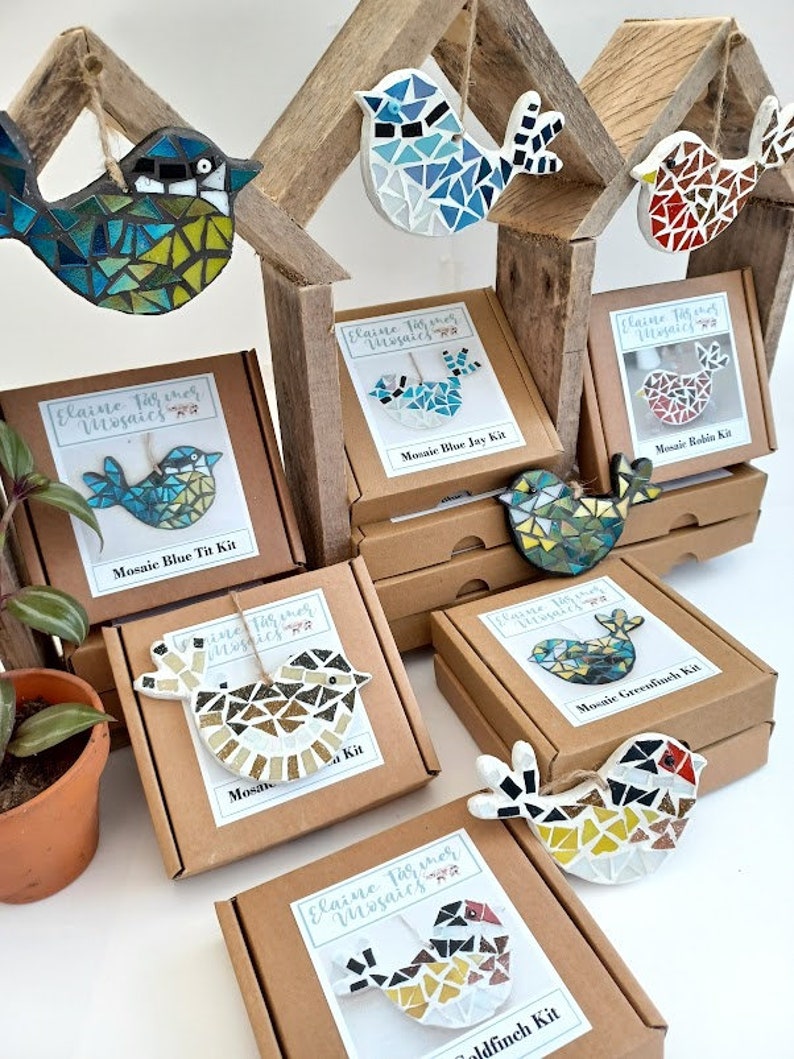 May include: A collection of mosaic bird kits and finished bird ornaments. The kits are in brown boxes with the text "Elaine Farmer Mosaics". The mosaic birds are in various colors, including blue, green, red, and gold. The finished birds are hanging from a wooden structure.