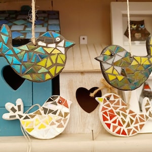 May include: Four mosaic bird ornaments in various colors, hanging from twine. The birds are made of small, colorful tiles, with blue, yellow, white, and red hues. They are displayed in front of a blue birdhouse and a white wooden structure.
