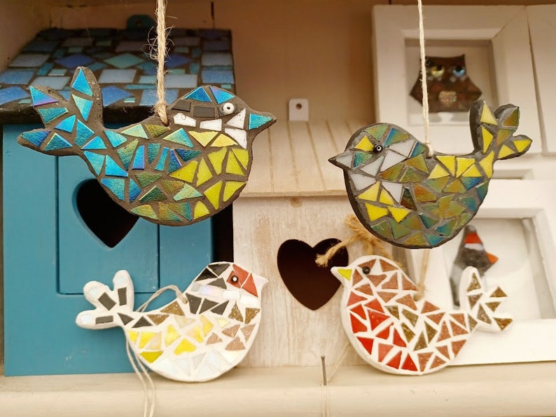 May include: Four mosaic bird ornaments in various colors, hanging from twine. The birds are made of small, colorful tiles, with blue, yellow, white, and red hues. They are displayed in front of a blue birdhouse and a white wooden structure.
