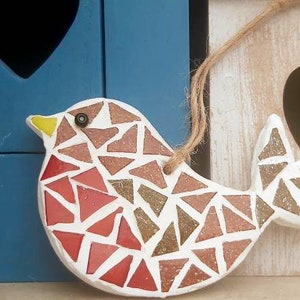 May include: A mosaic bird ornament with red, brown, and white triangular tiles. The bird is hanging from a string and has a yellow beak and black eye.