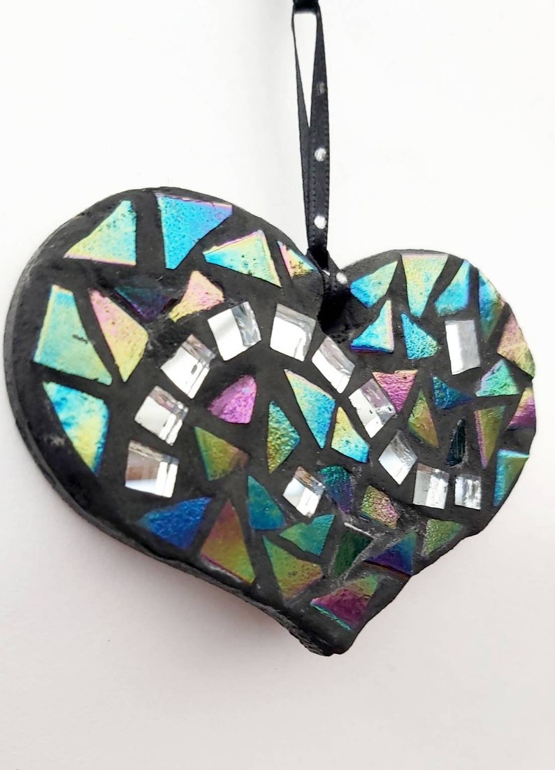 May include: A black heart-shaped ornament with a mosaic design. The mosaic is made of iridescent glass tiles in shades of blue, green, and purple. The ornament has a black ribbon for hanging.