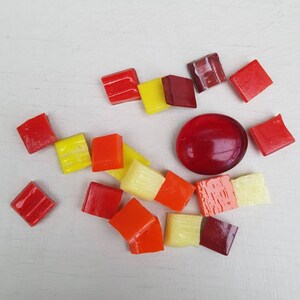May include: Assortment of small, colorful glass tiles in shades of red, orange, yellow, and cream. The tiles are arranged in a circular pattern on a white surface.