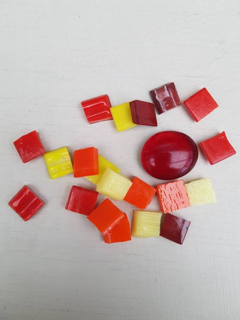 May include: Assortment of small, colorful glass tiles in shades of red, orange, yellow, and cream. The tiles are arranged in a circular pattern on a white surface.