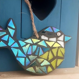 May include: A mosaic bird ornament with iridescent blue, green, and yellow glass pieces. The bird has a black outline and a jute string for hanging. The ornament is displayed in front of a blue birdhouse.