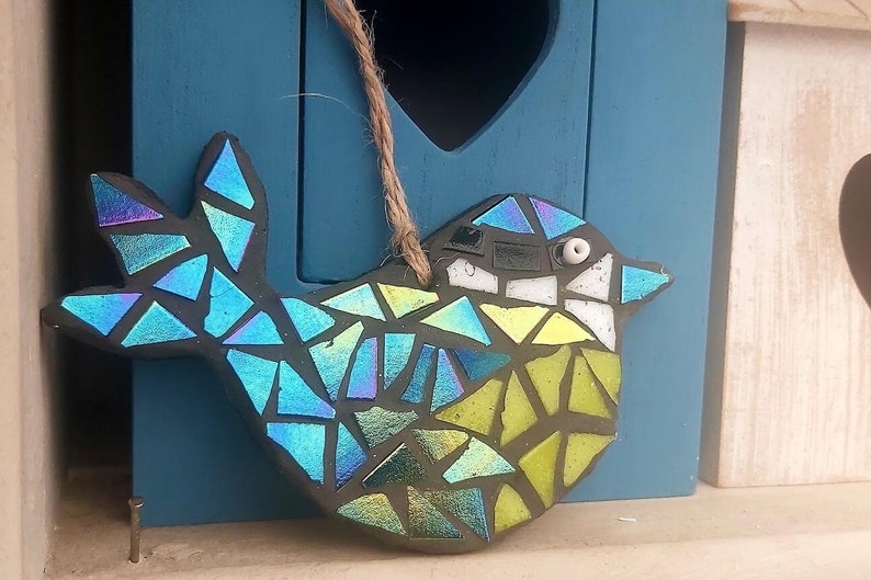 May include: A mosaic bird ornament with iridescent blue, green, and yellow glass pieces. The bird has a black outline and a jute string for hanging. The ornament is displayed in front of a blue birdhouse.