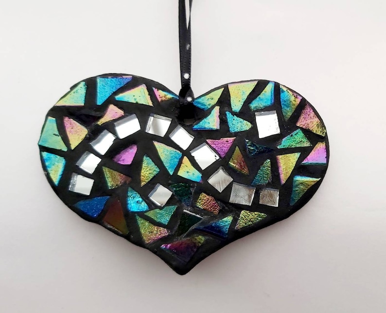 May include: A black heart-shaped ornament with a mosaic design made of iridescent glass tiles. The tiles are arranged in a random pattern and create a colorful and eye-catching effect.