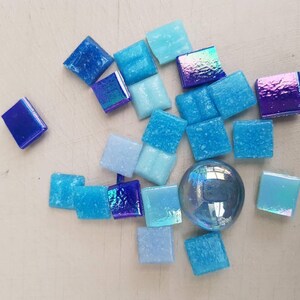 May include: Assortment of blue and iridescent glass mosaic tiles in various shades of blue, including light blue, dark blue, and iridescent blue. The tiles are arranged in a random pattern on a white surface.