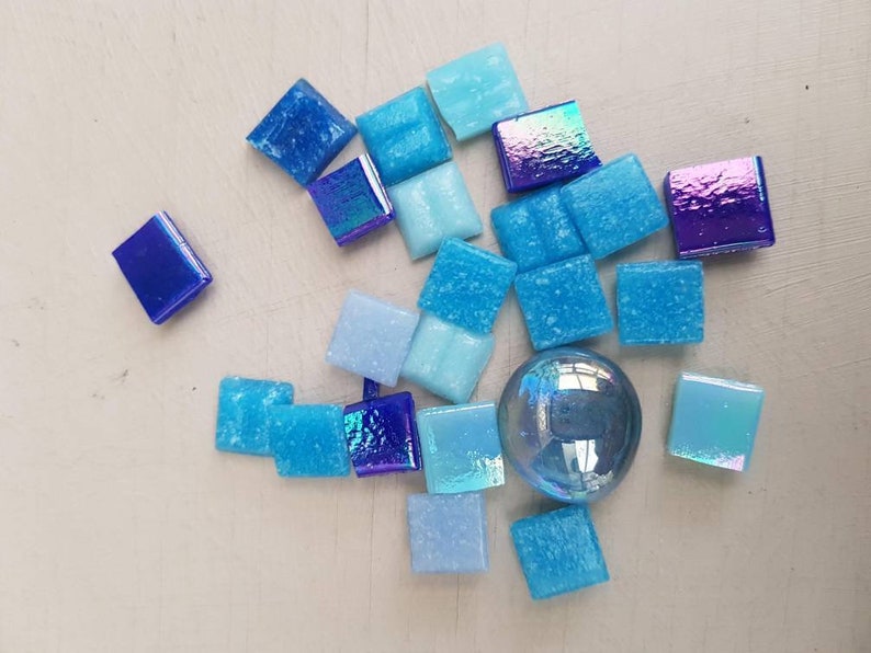 May include: Assortment of blue and iridescent glass mosaic tiles in various shades of blue, including light blue, dark blue, and iridescent blue. The tiles are arranged in a random pattern on a white surface.