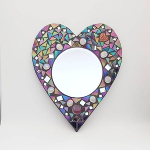 May include: A heart-shaped mirror with a mosaic frame made of iridescent glass tiles. The frame features a variety of colors, including pink, purple, blue, and green. A small red heart is in the center of the frame.