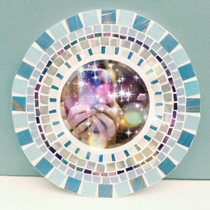 May include: A round mirror with a mosaic frame made of blue, white, and gray tiles. The mirror reflects a blurry image of a person holding a phone with a sparkling background.