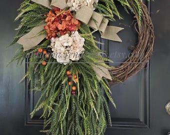 May include: A decorative wreath featuring a natural grapevine base adorned with cascading green fern-like foliage, cream and rust-colored hydrangea blossoms, small orange and dark purple berries, and a wide tan burlap ribbon tied in a bow.