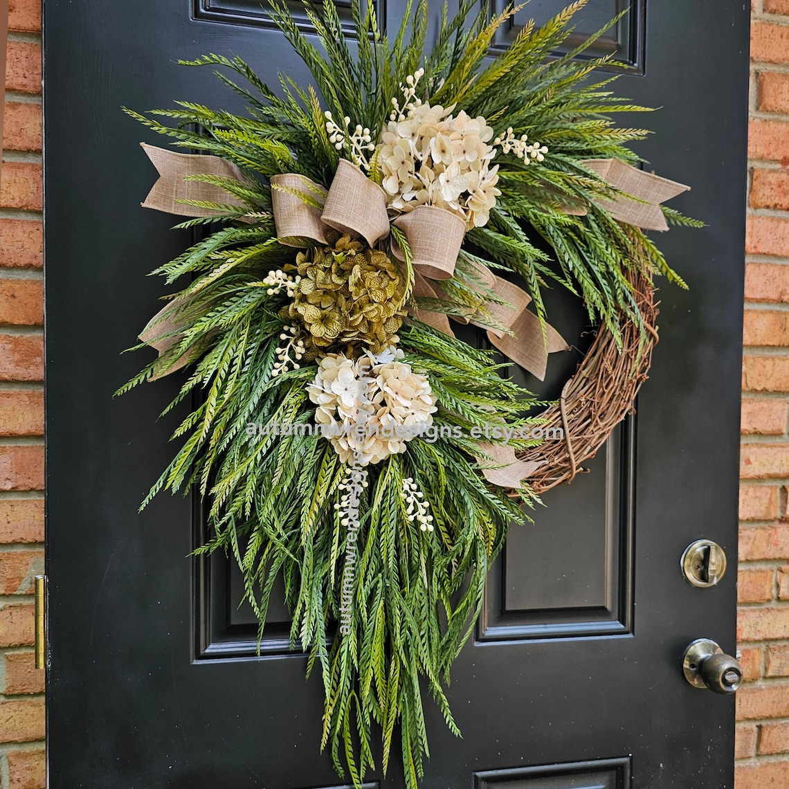 May include: A green and brown wreath with a burlap bow and white and green hydrangea flowers. The wreath is hanging on a black door.
