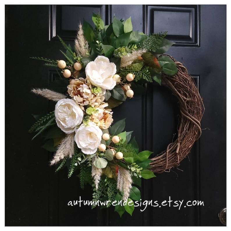 May include: A brown grapevine wreath decorated with white and beige flowers, green leaves, and beige dried grasses. The wreath is hanging on a black door with the text 'autumnwrendesigns.etsy.com' at the bottom.