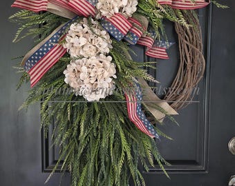 May include: A patriotic wreath with a grapevine base, adorned with green foliage, cream-colored flowers, and ribbons featuring the American flag. The wreath is designed for door decor.