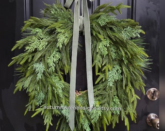 Op de afbeelding: Een weelderige, groene kunstkrans met een verscheidenheid aan kunstmatige groenblijvende bladeren. De krans is versierd met een saliegroen fluwelen lint om op te hangen. De krans wordt op een zwarte deur getoond. Tekst op de krans: autumnwrendesigns.etsy.com.