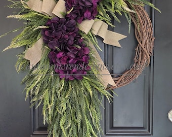 May include: A teardrop-shaped wreath featuring deep purple hydrangea blossoms, long green fern-like foliage, and a tan burlap ribbon. The arrangement is constructed on a circular brown twig base, creating a cascading botanical display.